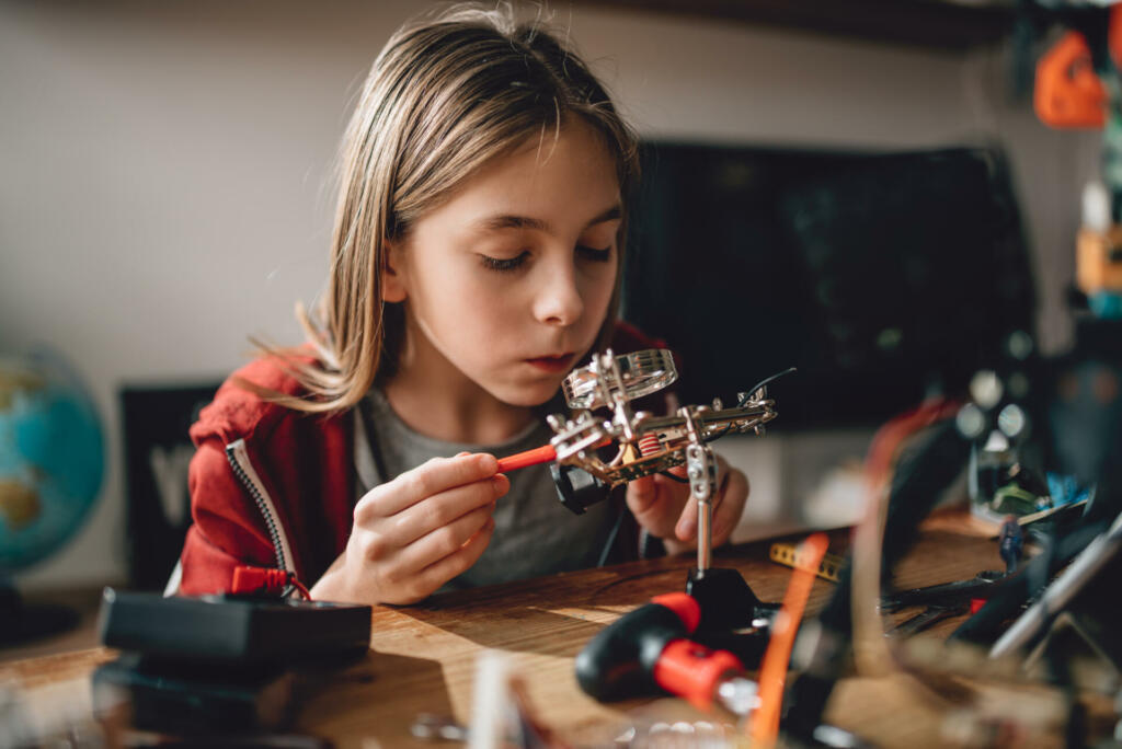 Girl wearing red hoodie looking circuit board throughout magnifying glass at home and building a robot