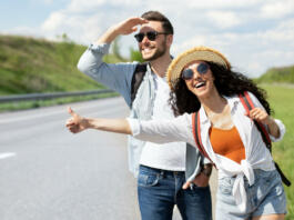 Happy young coupe flagging down car, needing ride, hitchhiking on roadside outdoors. Funky Caucasian man and his girlfriend traveling on summer vacation, having autostop journey