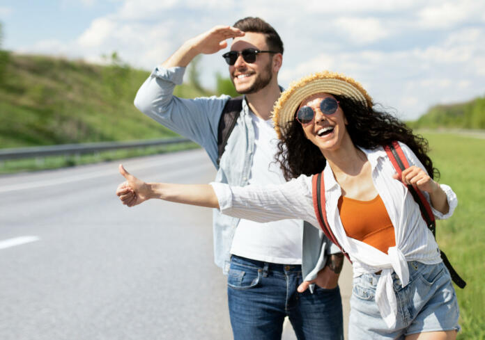Happy young coupe flagging down car, needing ride, hitchhiking on roadside outdoors. Funky Caucasian man and his girlfriend traveling on summer vacation, having autostop journey