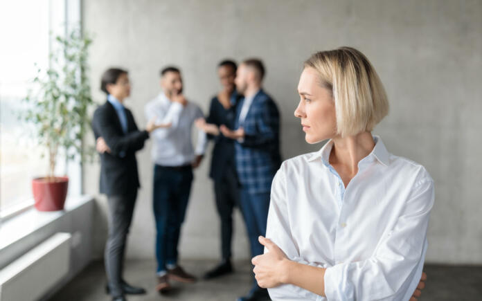 Male Coworkers Whispering Behind Back Of Unhappy Businesswoman Spreading Rumors And Gossips Standing In Modern Office. Sexism And Bullying Problem At Workplace Concept. Selective Focus