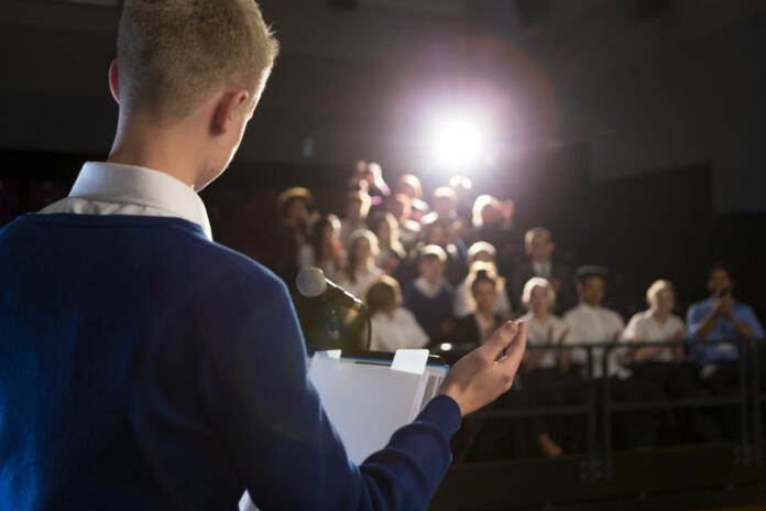 Male student making a speech. He is bstanding at the podium and is talking to the crowd of people.