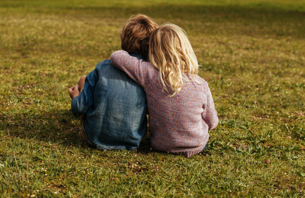 Rear shot of siblings sitting on the grass. Little girl sitting with her brother putting her arm on his shoulder at park.