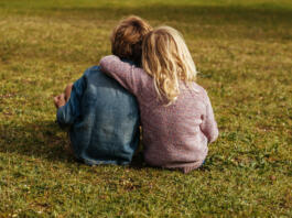 Rear shot of siblings sitting on the grass. Little girl sitting with her brother putting her arm on his shoulder at park.