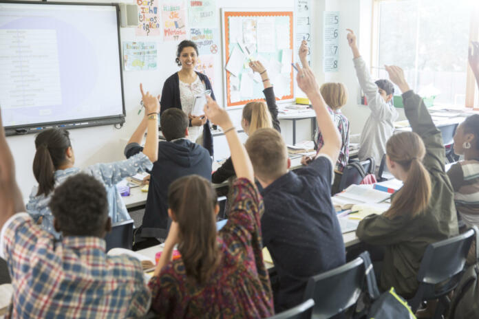 Rear view of teenage students raising hands in classroom