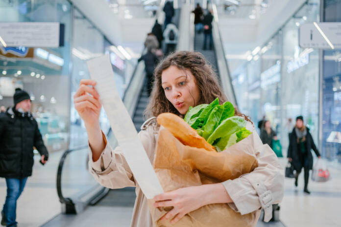 Rising food prices. Surprised woman looking into a paper check at the mall, holding a paper bag with fresh herbs and a baguette.