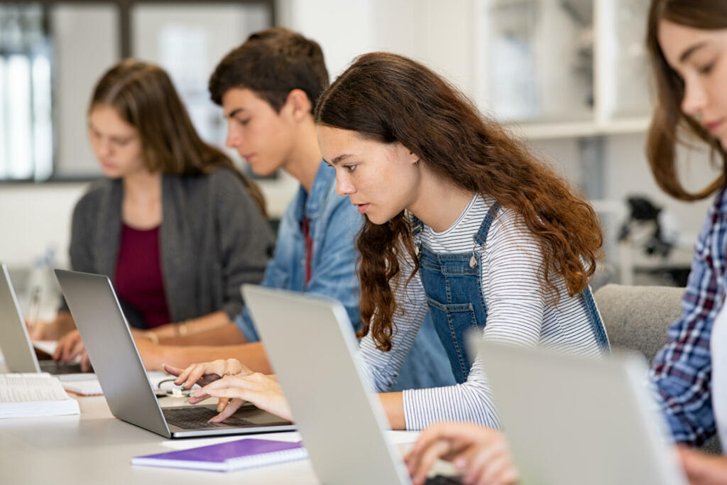 Serious college students studying on laptop sitting in a row in library. Young university multiethnic students using computer for study in classroom. Side view of casual girl typing on laptop during computer lesson.