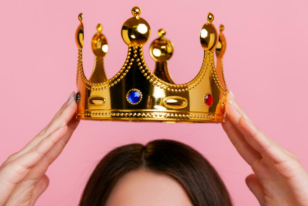 Unknown woman putting on golden crown, arrogance and privileged status, concept of self confidence in success, self-motivation and dreams to be best. Indoor studio shot isolated on pink background.