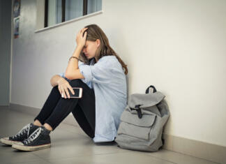 Duševna stiska mladih: kam po pomoč? Upset and depressed girl holding smartphone sitting on college campus floor holding head. University sad student suffering from depression sitting on floor at high school. Lonely bullied teen in difficulty with copy space.