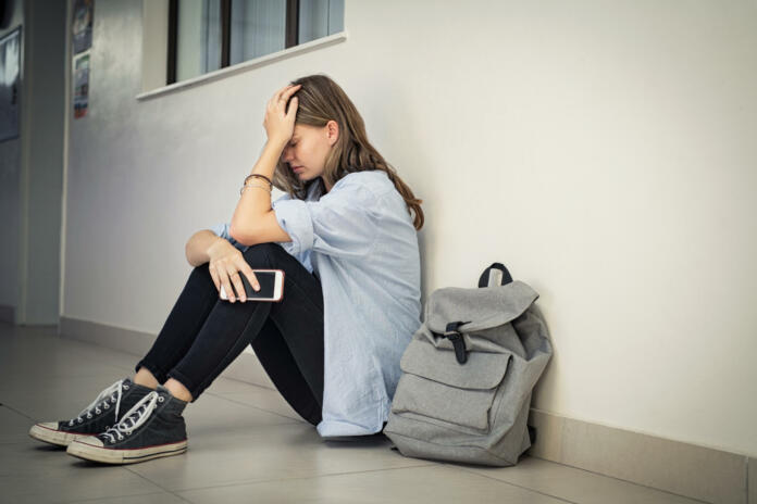 Upset and depressed girl holding smartphone sitting on college campus floor holding head. University sad student suffering from depression sitting on floor at high school. Lonely bullied teen in difficulty with copy space.