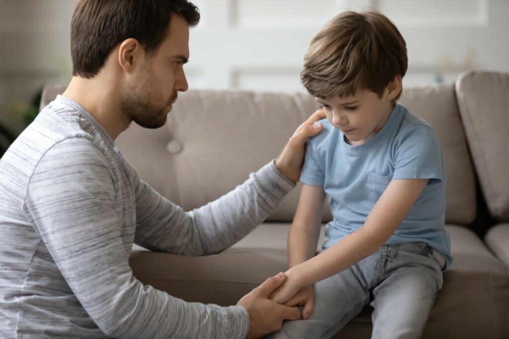 Worried young father holding hand of stressed little kid son, giving psychological help at home. Compassionate kind daddy supporting depressed offended small boy suffering from school bullying.