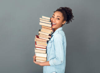 7 načinov, kako si med branjem knjig zapomniti več Young african female student isolated on grey wall holding a pile of books standing profile looking camera laughing