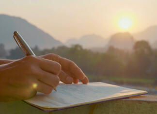 Navada, ki ti lahko korenito spremeni življenje Young woman writing travel notes in diary during sunrise with beautiful sun light and mountain landscape on the background, close-up.