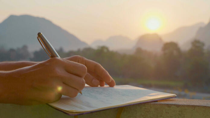 Young woman writing travel notes in diary during sunrise with beautiful sun light and mountain landscape on the background, close-up.