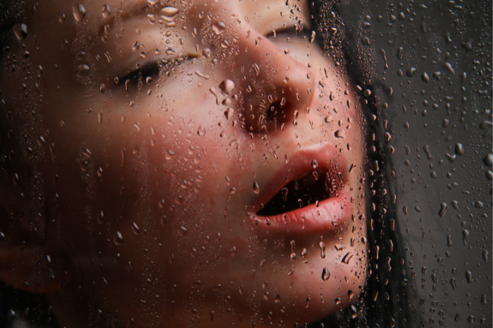 Ženska z zaprtimi očmi in odprtimi usti za stekleno steno, na kateri so vidne kapljice