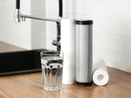 A glass of clean fresh water and set of filter cartridges on wooden table in a kitchen interior. Installation of reverse osmosis water purification system. Concept Household filtration system