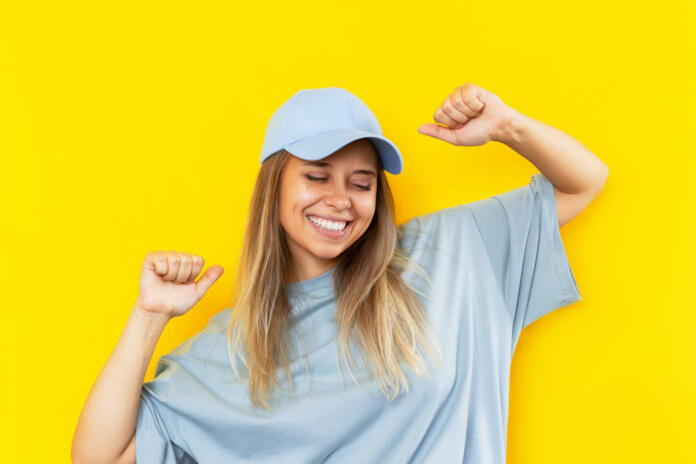 A young pretty caucasian excited smiling blonde woman in a grey t-shirt and blue cap is happy about the news or lottery win isolated on color yellow background. Cheerful girl dances with her hands up