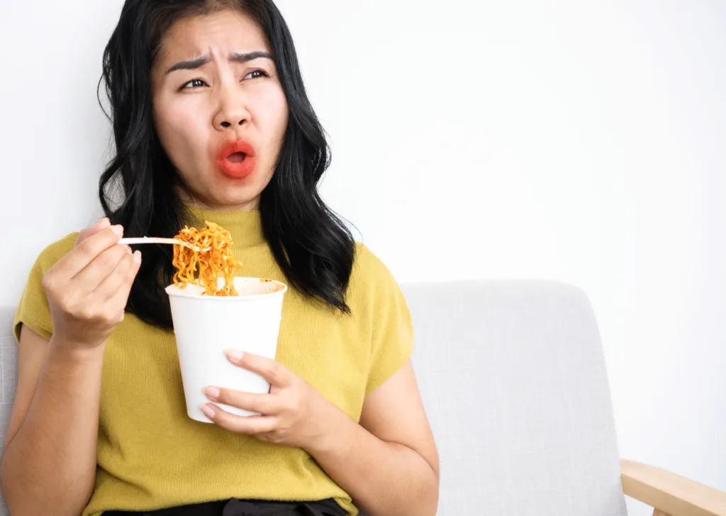 Asian woman eating very hot and spicy noodle from a cup her mouth and tongue burning and red, unhealthy eating concept
