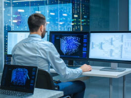 Beautiful Male Computer Engineer and Scientists Create Neural Network at His Workstation. Office is Full of Displays Showing 3D Representations of Neural Networks.