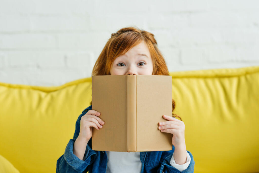 Child sitting on sofa and holding book in front of her face