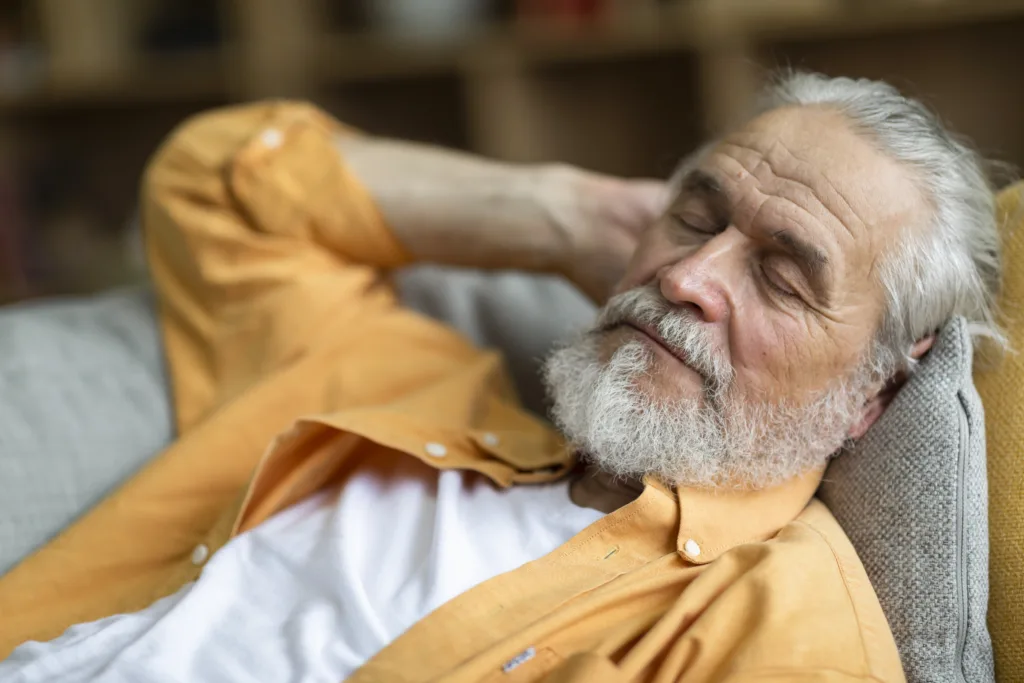 Closeup photo of sleeping senior man with long beard, peaceful grandfather resting on sofa at home, enjoying midday nap in living room. Seniors lifestyle concept