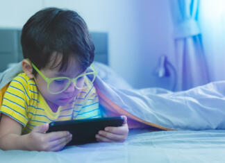 Se splača kupiti očala za modro svetlobo? Cute little child watch movie on smartphone at bed. Dangers of blue light can damage eyes. Handsome little boy can be age related macular degeneration from blue light, wear eyeglasses since childhood