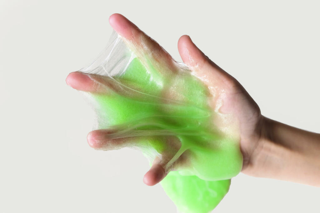 Hand Playing with textured slime on white background. Teen hand holding green shining slime with bubbles, stretching gooey substance. Teenage Girl squeezing slime toy to the sides. Liquid toy. Handgum. Top View, Flat Lay, Copy Space. View from Above. Horizontal banner. glurch, glorp. silly putty