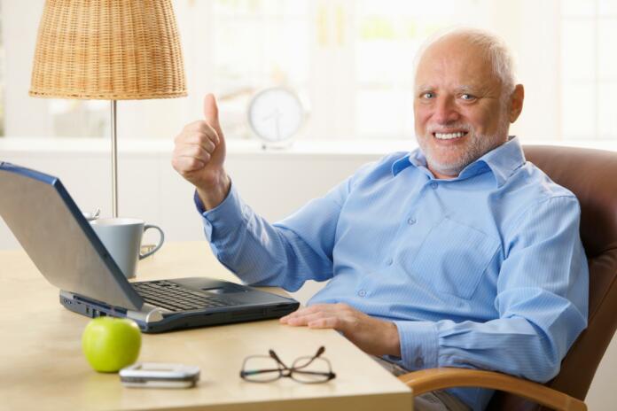 Happy senior man giving thumb up, sitting at desk using laptop computer at home...