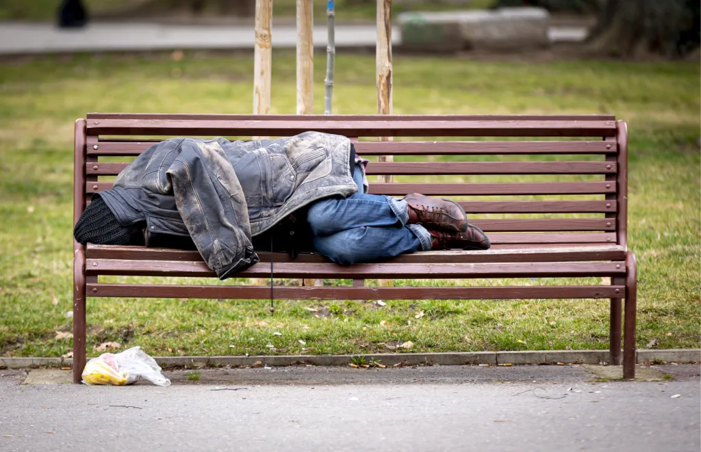 Homeless man sleeps on a bench covered with his jacket in a public park during the day.