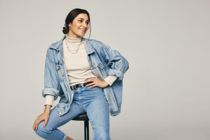 Rocking denim wear. Fashionable young woman sitting on a chair against a grey background. Happy young woman looking away with a smile on her face in a modern studio.