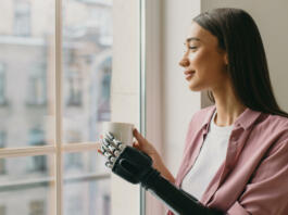 Side view of beautiful lovely female with long brown hair standing in front of window with coffee in hands, watching her boyfriend returning home, having iron biotechnological arm prosthesis