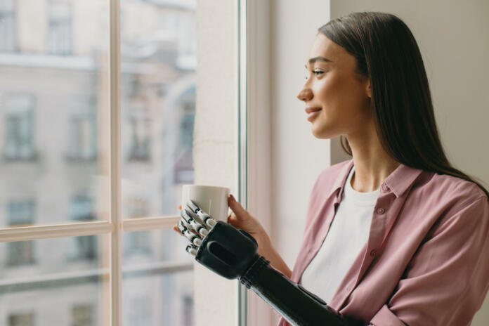 Side view of beautiful lovely female with long brown hair standing in front of window with coffee in hands, watching her boyfriend returning home, having iron biotechnological arm prosthesis