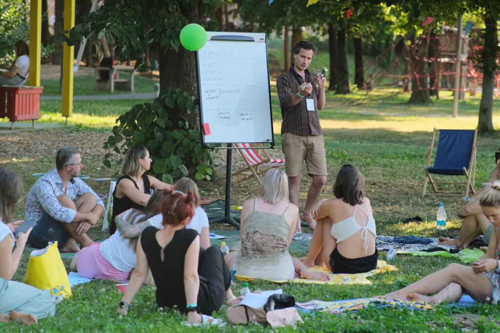 Udeleženci festivala sedijo na tleh pred predavateljem, ki stoji pred tablo