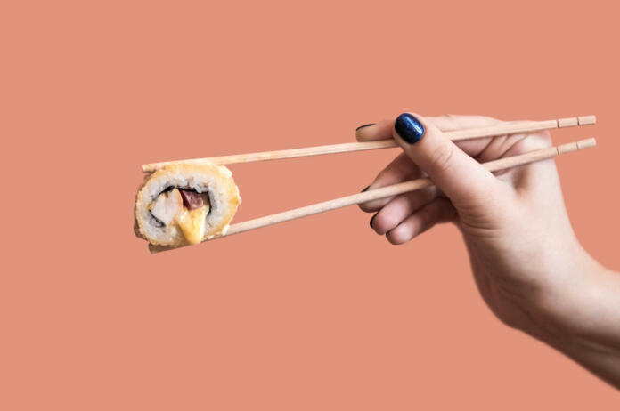 A hand holds a fried sushi roll with cheese with wooden sticks.