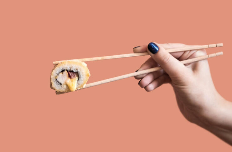 A hand holds a fried sushi roll with cheese with wooden sticks.