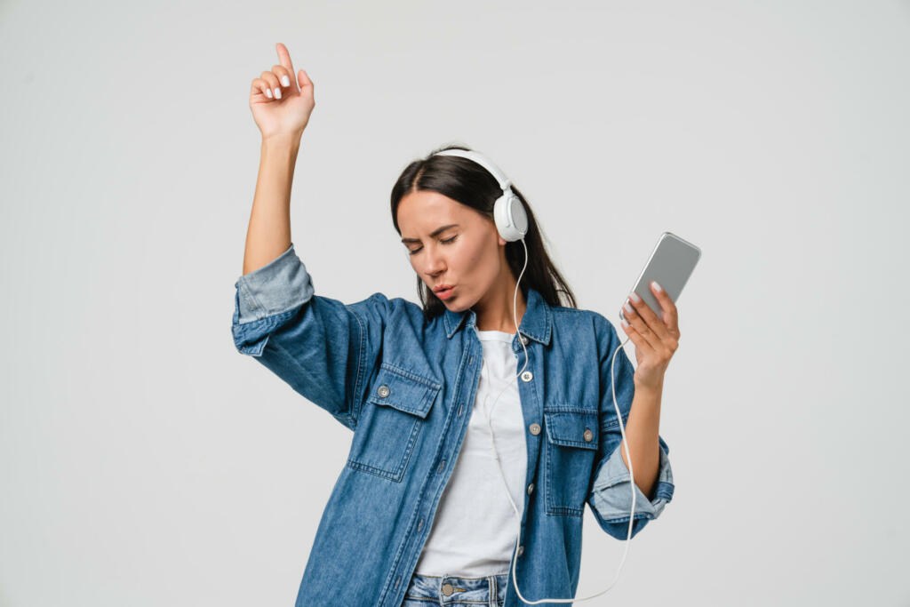 Energetic caucasian young woman girl dancing singing listening to the music podcast song singer sound track e-book in headphones earphones on cellphone isolated in white background