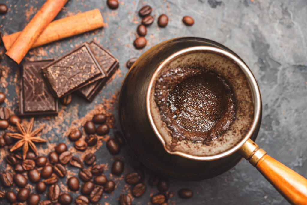 Fresh brewed coffee in the turkish jezva coffee pot on dark background with coffee beans and chocolate pieces, top view.