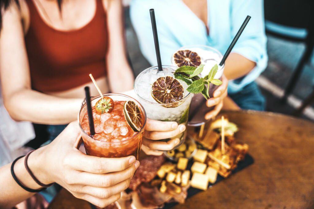 Happy friends clink cocktail glasses at bar restaurant - Young people enjoying happy hour toasting drinks at outside dinner party - Food and beverage life style concept with girls drinking outdoors