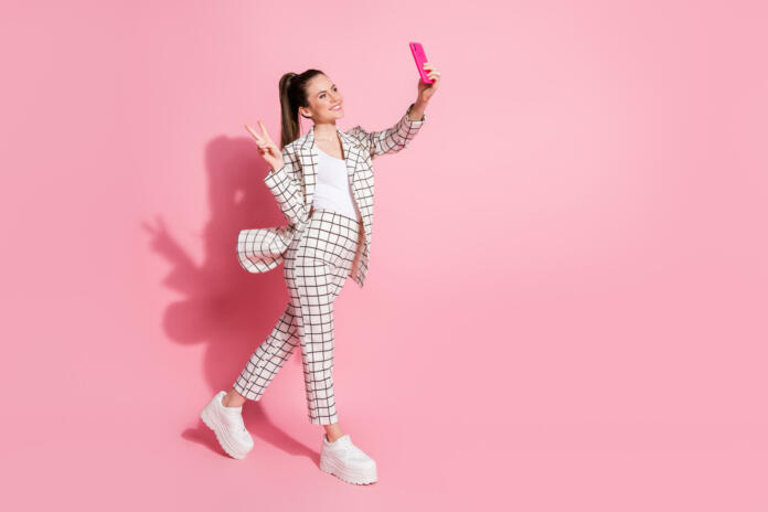 Photo portrait full body view of woman taking selfies on the go showing v-sign isolated on pastel pink colored background.