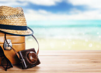 Kako potovati bolj trajnostno? Vse o zelenem potovanju Vintage suitcase, sunglasses and hipster hat wooden deck and blur tropic sea background. Summer paradise cruise travel design layout banner. Tourism, beach, vacation travel destination concept wallpaper mockup.