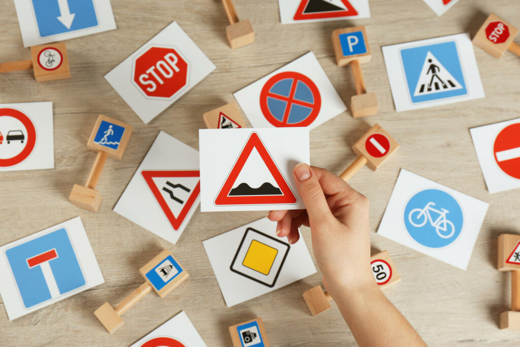 Woman holding rough road sign over wooden table, top view. Driving license exam