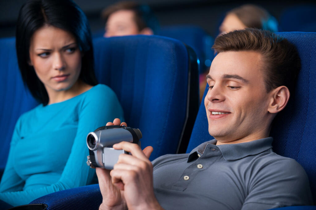 Young man shooting with his home video camera while sitting close to the woman at the cinema