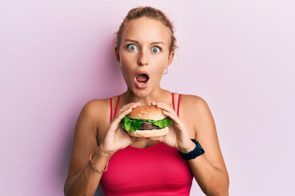 Beautiful caucasian woman eating a tasty classic burger afraid and shocked with surprise and amazed expression, fear and excited face.