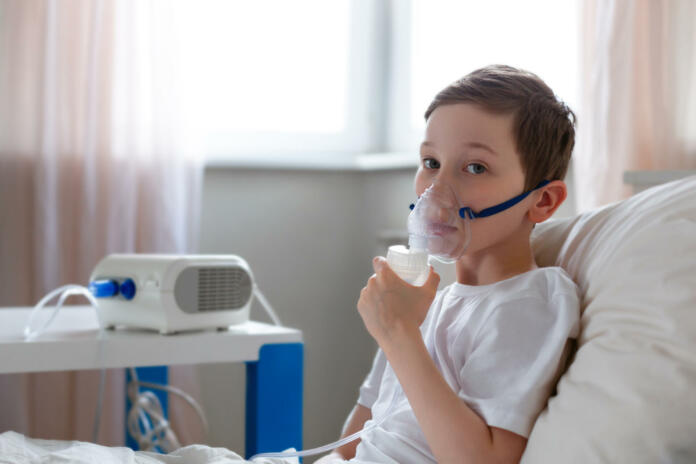 Cute boy inhalation therapy by the mask of inhaler. Kid with respiratory problem or asthma with copy space. View of portable nebulizer with smoke from oxygen mask.
