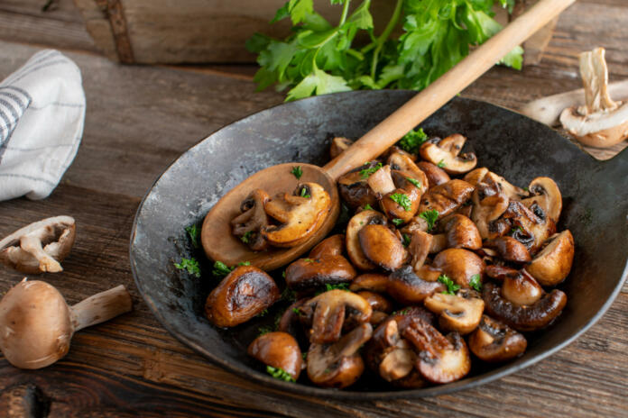 Delicious side dish or snack with pan fried mushrooms, champignons cooked with butter, herbs, pepper and salt. Served in a rustic cast iron pan on wooden table.