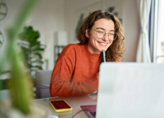 8 nasvetov za sestanek na daljavo Happy young woman using laptop sitting at desk writing notes while watching webinar, studying online, looking at pc screen learning web classes or having virtual call meeting remote working from home.