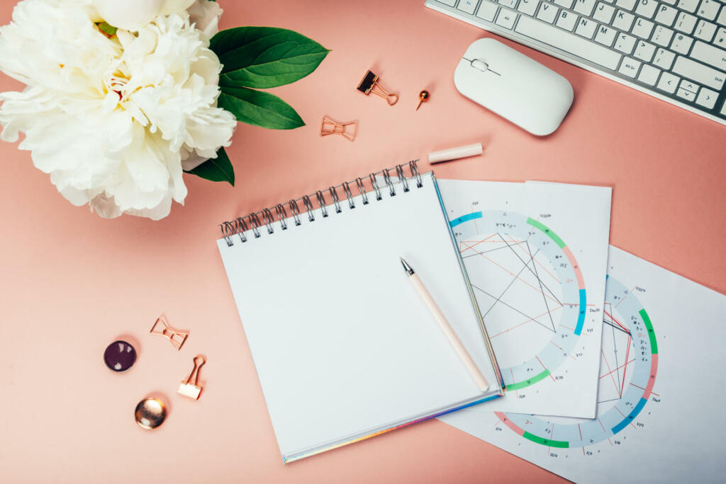 Open Notebook for writing ideas and astrological birth natal chart laying on pink background. Computer keyboard and mouse nearby. Idea of Astrological Consultation. Flat lay in girly style