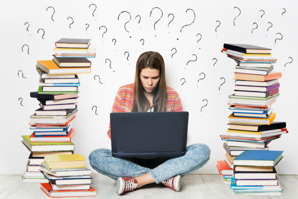 Woman using Laptop. Student Girl working on Computer Search in Internet around Stacks of Books. Question Marks Drawn On White Wall Background. E Learning, Online Studying, Reading Books in Digital Library. Young Girl sitting on Floor next to heap of Books