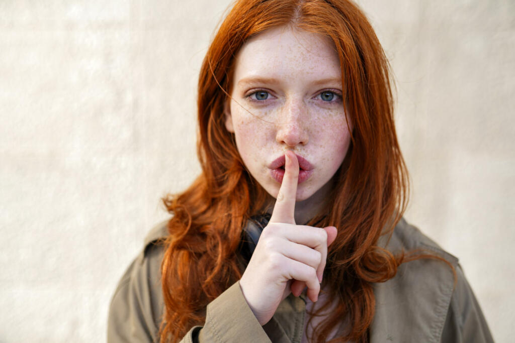 Hipster teen gen z redhead girl showing shh sign finger gesture asking to keep secret, be hush silent or privacy silence on urban wall background. Teenage problem secrecy concept. Close up portrait