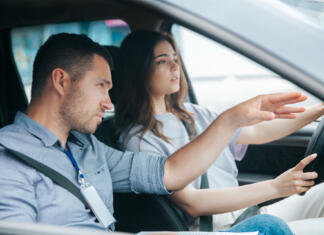10 najpogostejših napak na izpitni vožnji in nasveti Male instructor showing something by his hand on the road and giving advice. Attractive woman listening her teacher, looking ahead with concentration and holding wheel