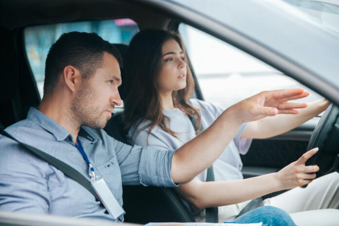 Male instructor showing something by his hand on the road and giving advice. Attractive woman listening her teacher, looking ahead with concentration and holding wheel